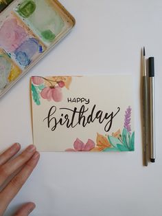 a hand holding a card with the words happy birthday on it next to some watercolor paints