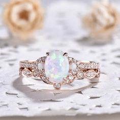 a white opal and diamond ring on top of a lace doily with flowers in the background