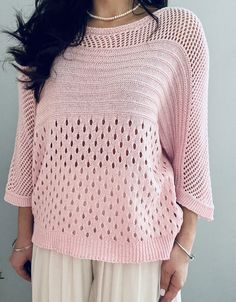 a woman standing in front of a wall wearing a pink knitted sweater and white pants