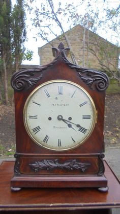 an old clock sitting on top of a wooden table in front of a tree and building