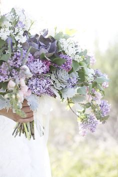 a woman holding a bouquet of purple and white flowers with greenery in her hands