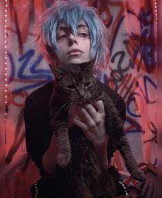 a woman with blue hair holding a cat in front of a wall covered in graffiti