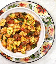 a white bowl filled with pasta and meat on top of a colorful table cloth next to a spoon