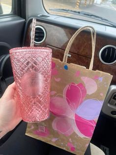 a hand holding a pink glass in front of a brown paper bag with flowers on it