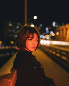 a woman standing on the side of a road at night with lights in the background