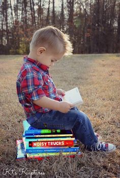 a young boy sitting on top of a pile of books in the grass with trees behind him