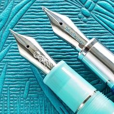 two fountain pens sitting next to each other on a blue towel with silver trimmings