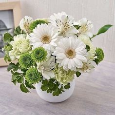 a vase filled with white and green flowers on top of a wooden table next to a framed photo