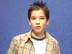 a young man standing in front of a blue wall wearing a plaid shirt and looking at the camera