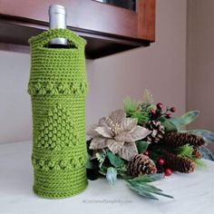 a green knitted water bottle cover next to pine cones and greenery on a counter