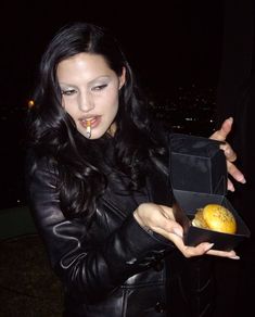 a woman in black leather jacket holding up a box with food on it and looking at the camera