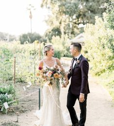 a bride and groom holding hands walking down a path in the middle of a garden