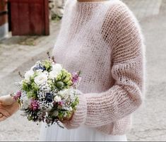 a woman in a white dress holding a bouquet of flowers and wearing a pink sweater