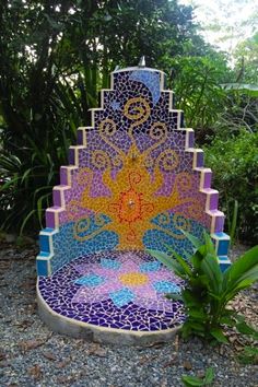 a bench made out of mosaic tiles in the middle of some bushes and trees with plants around it