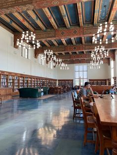 people are sitting at long tables in the middle of a large room with bookshelves and chandeliers