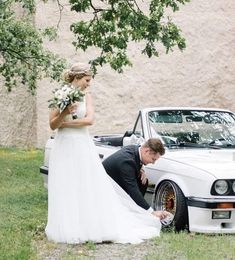 a bride and groom standing in front of a white car with their hands on the ground