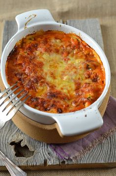 a white casserole dish with cheese and tomato sauce on a wooden board next to a fork