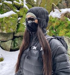 a woman with long hair wearing a black mask and jacket standing in front of snow covered rocks