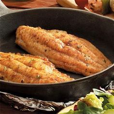 two fish fillets in a frying pan on a table with vegetables and bread