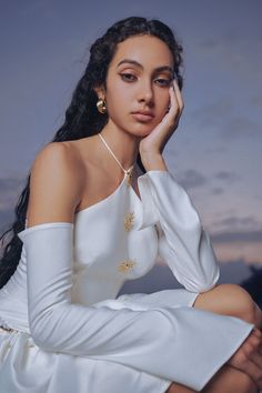 a woman in a white dress sitting on the ground with her hand under her chin