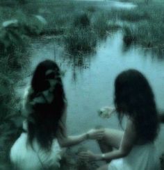 two women sitting on the ground in front of a body of water with grass and plants