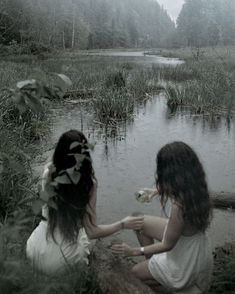 two young women sitting on the ground next to a body of water, eating food