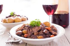 a white bowl filled with meat and potatoes next to two glasses of wine