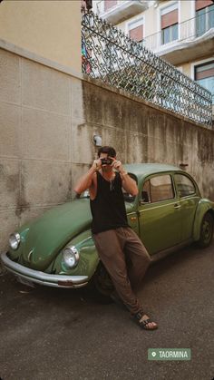 a man leaning on the hood of a green car while taking a photo with his cell phone