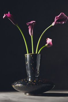 three pink flowers in a black vase on a gray tablecloth, with dark background