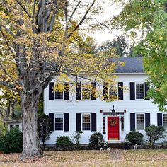 a white house with black shutters and red door in the front yard surrounded by trees