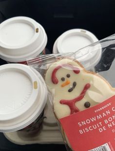 there is a frosted snowman cookie in the plastic wrapper next to some coffee cups