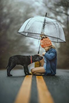 a woman kneeling down next to a black dog on the side of a road holding an umbrella