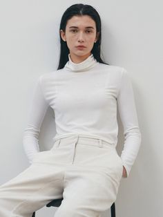 a woman sitting on top of a chair wearing white pants and a turtle neck sweater