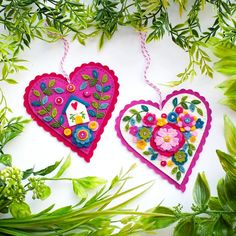 two heart shaped ornaments hanging from green leaves on a white surface with flowers in the background