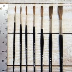 six brushes are lined up next to a ruler on a wooden surface, with one being used as an eyeliner brush