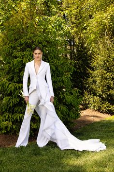 a woman in a white suit and dress standing on grass with trees in the background