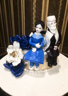 a cake with two figurines sitting on it's side, one holding a flower and the other wearing a blue dress