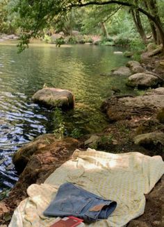 an open book is laying on a blanket near the water's edge, with rocks and trees in the background