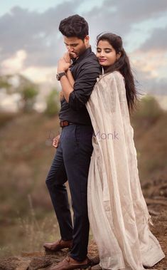 a man and woman standing next to each other on top of a dirt hill with clouds in the background