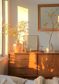 the sun shines through two windows onto a dresser with vases and flowers on it