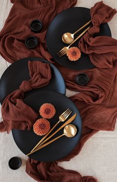 two black plates with gold forks and spoons on top of a brown table cloth