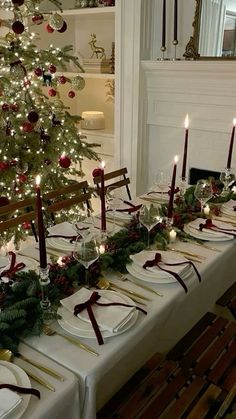 a table set for christmas dinner with lit candles and plates on it, next to a decorated christmas tree