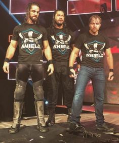 three men in black shirts standing on stage with one man wearing knee pads and boots