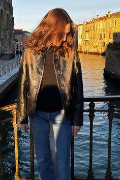 a woman standing on a bridge next to the water wearing a black jacket and jeans