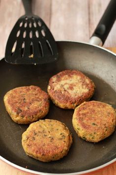 four crab cakes cooking in a frying pan on a wooden table next to a spatula