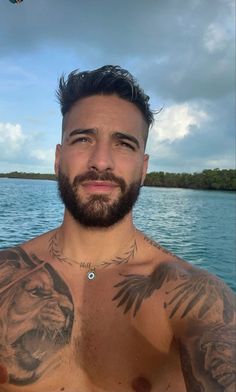 a man with tattoos on his chest standing in front of water