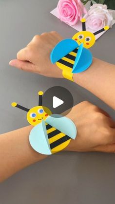 two people wearing paper bee bracelets with flowers in the background and one person's hand holding a flower