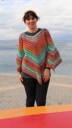 a woman standing next to the ocean wearing a multicolored crocheted sweater