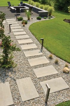 a stone path in the middle of a garden with grass and bushes around it, leading to an outdoor seating area