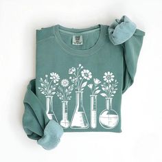 Embrace the cozy side of science with our Sleek Beaker Botanical Sweatshirt. This unique pullover showcases a stunning design of delicate flowers emerging from various laboratory beakers and flasks, perfectly capturing the intersection of botany and chemistry. Ideal for chilly days in the lab or classroom, this sweatshirt offers both warmth and style to science teachers, chemists, and biology enthusiasts alike. The soothing sage green color complements the intricate white floral and glassware design, creating a piece that's both sophisticated and fun. Whether you're conducting experiments, grading papers, or simply enjoying a casual day out, this sweatshirt is sure to spark curiosity and admiration from fellow science lovers. It's the perfect way to wear your passion for science and nature Biology Lab, Biology Labs, Glassware Design, Lab Technician, Sage Green Color, Science Lover, Beakers, Floral Sweater, Color Complement